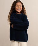 A smiling child with long curly hair, wearing the navy blue Kids Cotton Fisherman sweater and cream pants, stands with arms crossed against a plain beige background.