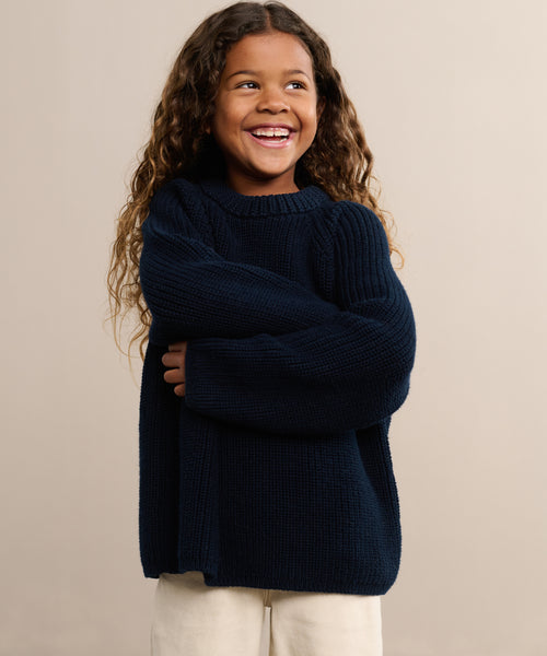 A smiling child with long curly hair, wearing the navy blue Kids Cotton Fisherman sweater and cream pants, stands with arms crossed against a plain beige background.