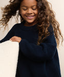 Smiling young girl with long curly hair wears a dark blue Kids Cotton Fisherman sweater and holds a cream-colored textured object, standing against a light background.