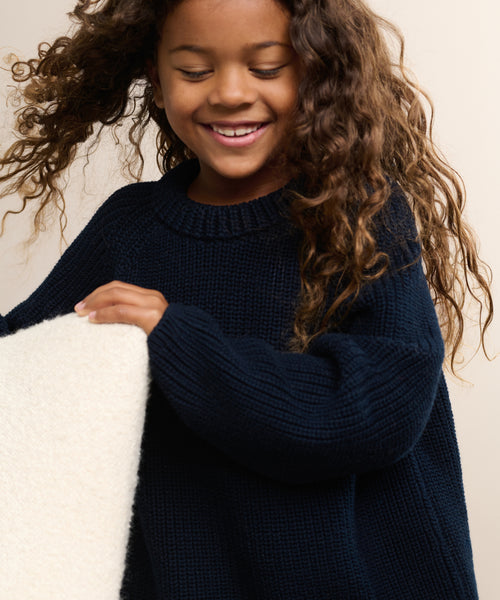 Smiling young girl with long curly hair wears a dark blue Kids Cotton Fisherman sweater and holds a cream-colored textured object, standing against a light background.