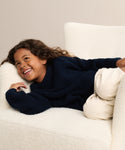 A young child with long curly hair, smiling, lies sideways on a white textured armchair, wearing the Kids Cotton Fisherman sweater in navy blue and light beige pants.
