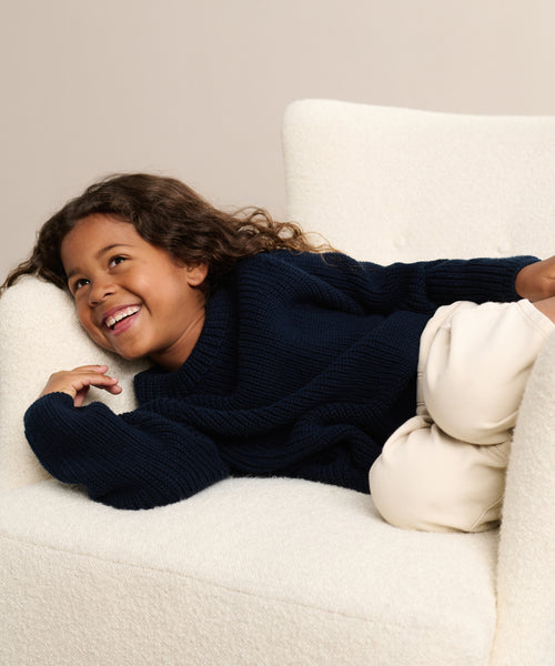 A young child with long curly hair, smiling, lies sideways on a white textured armchair, wearing the Kids Cotton Fisherman sweater in navy blue and light beige pants.