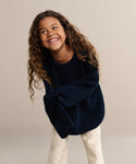 A young child with long curly hair smiles and leans forward against a plain beige background, wearing the Kids Cotton Fisherman sweater in dark blue paired with light-colored pants.