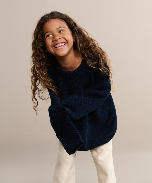 A young child with long curly hair smiles and leans forward against a plain beige background, wearing the Kids Cotton Fisherman sweater in dark blue paired with light-colored pants.