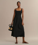A woman wears the Kinney Dress, a sleeveless black summer style, standing against a neutral background with a small woven handbag and simple sandals.