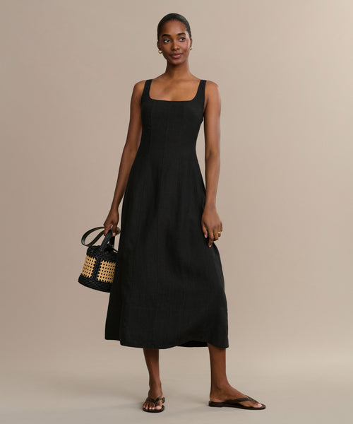 A woman wears the Kinney Dress, a sleeveless black summer style, standing against a neutral background with a small woven handbag and simple sandals.