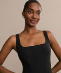 A woman with smooth dark skin and pulled-back hair softly smiles in a sleeveless Kinney Dress with a square neckline, standing before a plain beige background.