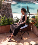 Wearing the Kinney Dress and flip-flops, a woman sits calmly on a metal chair by a sunlit pool, framed by stone walls and potted plants.