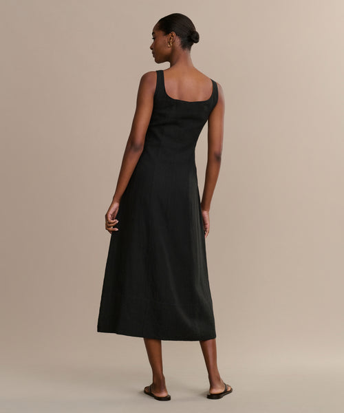A woman stands on a neutral background wearing the Kinney Dress, a sleeveless black summer dress with a square neckline, paired with flat sandals. She faces away, looking to the side, her hair styled in a neat bun.