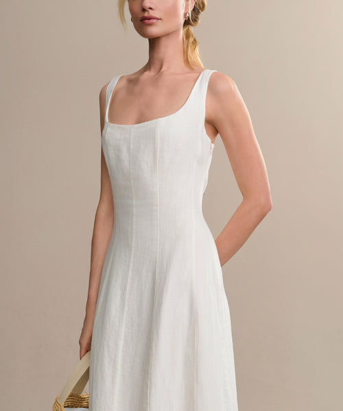 A woman in the Kinney Dress, a sleeveless white linen summer dress with a square neckline, stands against a plain beige background, holding a woven handbag by her side.