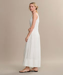 A woman stands sideways in the Kinney Dress, a long sleeveless linen summer dress, paired with flat sandals. Her arms are gently crossed and her hair is in a loose low bun, set against a plain beige background.