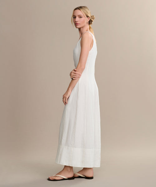 A woman stands sideways in the Kinney Dress, a long sleeveless linen summer dress, paired with flat sandals. Her arms are gently crossed and her hair is in a loose low bun, set against a plain beige background.