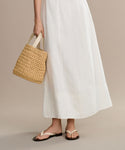 A person wearing the Kinney Dress and white sandals holds a woven straw handbag by their side against a plain, neutral background.