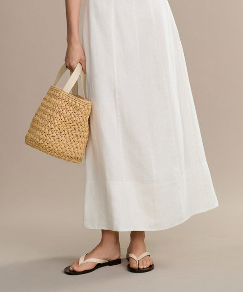A person wearing the Kinney Dress and white sandals holds a woven straw handbag by their side against a plain, neutral background.