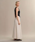 A woman with blonde hair stands sideways against a beige backdrop, wearing a sleeveless black top, black sandals, and the Kinney Skirt—a long, lightweight linen skirt ideal for summer. She has her hair styled back and a neutral expression.