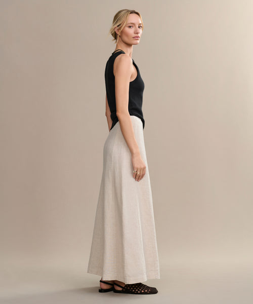 A woman with blonde hair stands sideways against a beige backdrop, wearing a sleeveless black top, black sandals, and the Kinney Skirt—a long, lightweight linen skirt ideal for summer. She has her hair styled back and a neutral expression.