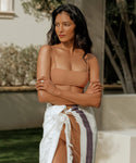 A woman with long dark hair stands outdoors, wearing a Kumu Bikini Top and a white towel with colorful stripes tied at her waist, arms crossed and looking to the side, with greenery and a building in the background.