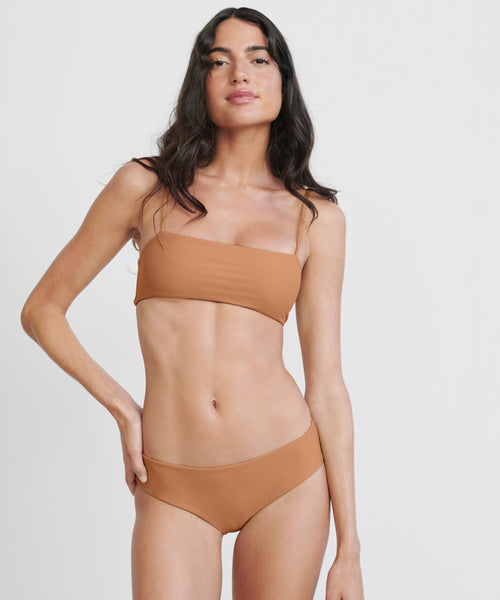 A woman with long dark hair wears the Kumu Bikini Top in terracotta, posing confidently against a plain white background.