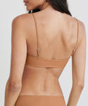 A woman with long dark hair is seen from behind, wearing a Kumu Bikini Top in tan with adjustable straps, set against a plain light background.