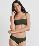 A woman with long brown hair poses against a plain light background, smiling softly as she touches her hair. She wears the Kumu Bikini Top by Mikoh in olive green, featuring thin straps and a straight neckline, made from recycled nylon.