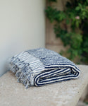 The Laguna Beach Towel, featuring blue and white stripes and fringed edges, is folded on a stone surface with greenery softly blurred in the background.