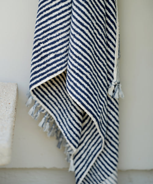 A close-up of the Laguna Beach Towel, showing navy blue and white diagonal stripes with fringed edges. Made from Turkish cotton, this towel looks soft and cozy, its textured fabric draped against a light background.
