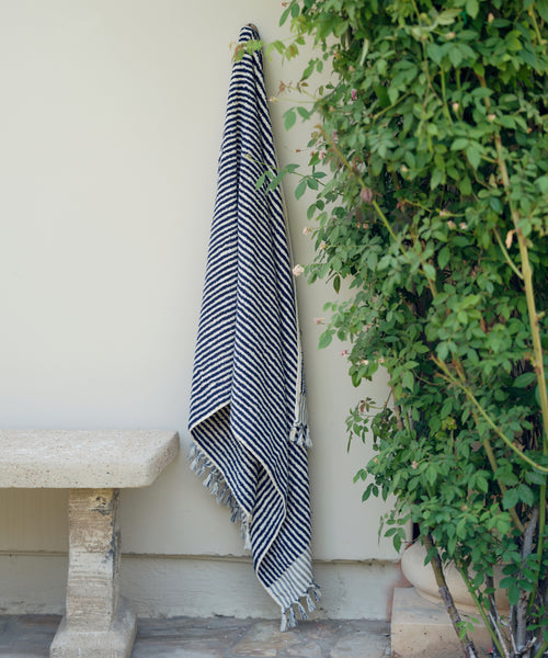 The Laguna Beach Towel, featuring black and white stripes and fringed edges, hangs on a light wall near a green leafy bush and a stone bench.