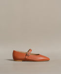 The Lana Flat is an orange-brown leather Mary Jane shoe featuring a tapered toe and a thin strap with a small buckle across the instep, shown on a neutral background.