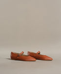 The Lana Flat is an orange-brown Mary Jane with thin straps, small buckles, and a tapered toe, shown on a light beige background.
