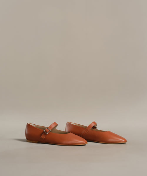 The Lana Flat is an orange-brown Mary Jane with thin straps, small buckles, and a tapered toe, shown on a light beige background.