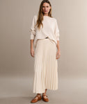 A woman with long, wavy hair poses casually against a beige background, wearing a loose cream sweater, a pleated cream skirt, and the Lana Flat—a brown Mary Jane shoe with a tapered toe that adds feminine flair.