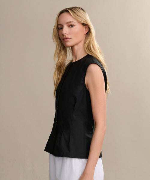 A woman with long blonde hair models the sleeveless black Lara Top and white pants, standing sideways against a plain beige background, gazing at the camera with a neutral expression.