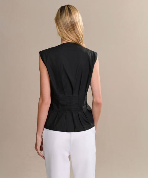 A woman with straight blonde hair, seen from behind, wears the black sleeveless Lara Top with pleats and white pants against a plain beige background.