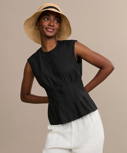 A woman models the Lara Top—a sleeveless black button-front shirt—paired with white pants and a wide-brimmed straw hat, standing against a neutral background with a relaxed smile and her arms slightly bent.