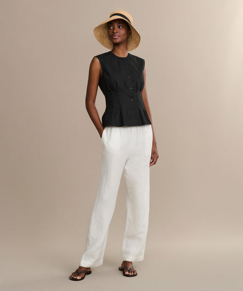 A woman wears the Lara Top—a sleeveless black button-front style—paired with white loose pants, black sandals, and a wide-brimmed straw hat, standing against a plain beige background.