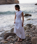 Wearing the sleeveless white Lara Top, made from Italian washed poplin, she stands in sandals on a rocky beach at sunset, gazing downward as waves and cliffs form a stunning backdrop.