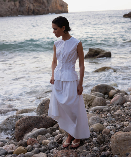 Wearing the sleeveless white Lara Top, made from Italian washed poplin, she stands in sandals on a rocky beach at sunset, gazing downward as waves and cliffs form a stunning backdrop.