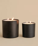 The Large Aspen Glass Candle, hand-poured in Los Angeles with three wicks, and a smaller black matte two-wick candle glow warmly atop a beige surface against a matching background.