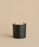 A black matte candle holder displays the Large Cedar Glass Candle (28oz, double wick), glowing warmly on a smooth beige surface against a matching background—an elegant piece inspired by Oprah’s Favorite Things.