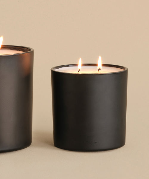 A Large Cedar Glass Candle, 28oz, in matte black with two wicks burns on a neutral surface; part of a similar jar appears on the left against a beige background—an elegant choice from Oprah’s Favorite Things.