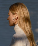 A woman with long blonde hair wears the Large Everyday Hoops, paired with a white turtleneck sweater, shown in profile against a blurred blue background—perfect lightweight earrings for daily wear.