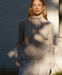 A woman with long blonde hair wears a cream cable-knit turtleneck, light pants, and Large Everyday Hoops outdoors. She stands in partial sunlight with soft shadows and a blurred building behind her—an effortlessly chic look.