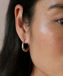 Close-up of a person’s ear with long dark hair, wearing Large Hellenic Hoops—thick gold vermeil earrings handcrafted in Los Angeles. The image highlights both the hoop and a portion of the wearer’s cheek and jawline.