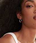 A close-up shows a woman with curly black hair wearing Large Sigrid Hoops in sterling silver and a white top. Only the right side of her face and shoulder are visible, featuring a beauty mark near her lip that highlights the handmade design.