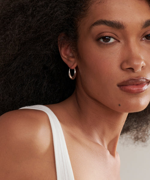 A woman with curly dark hair wears a white sleeveless top and handmade sterling silver Large Sigrid Hoops, gazing at the camera with a neutral expression.