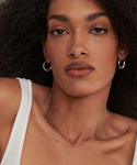 A person with natural curly hair, wearing Large Sigrid Hoops—sterling silver handmade jewelry—and a white top looks directly at the camera with a neutral expression.