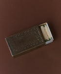 The Large Stamped Matchbox features a zigzag-patterned cover holding a white matchbox with green-tipped matches inside, shown partially open on a brown surface.