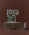 A Large Stamped Matchbox with a metallic cover and turquoise knob, inspired by Navajo silverwork, sits open to reveal matches inside, stacked against a brown background.