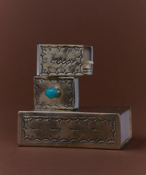 A Large Stamped Matchbox with a metallic cover and turquoise knob, inspired by Navajo silverwork, sits open to reveal matches inside, stacked against a brown background.