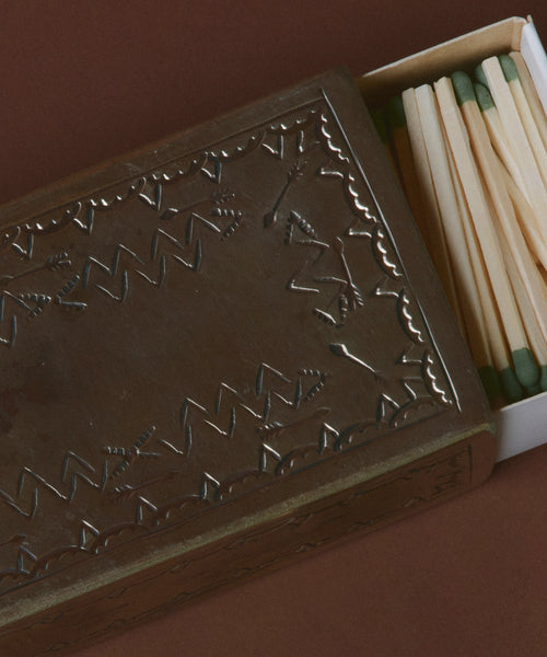 The Large Stamped Matchbox features an embossed zigzag and arrow design and is partially open to reveal green-tipped wooden matches inside, displayed against a brown background.